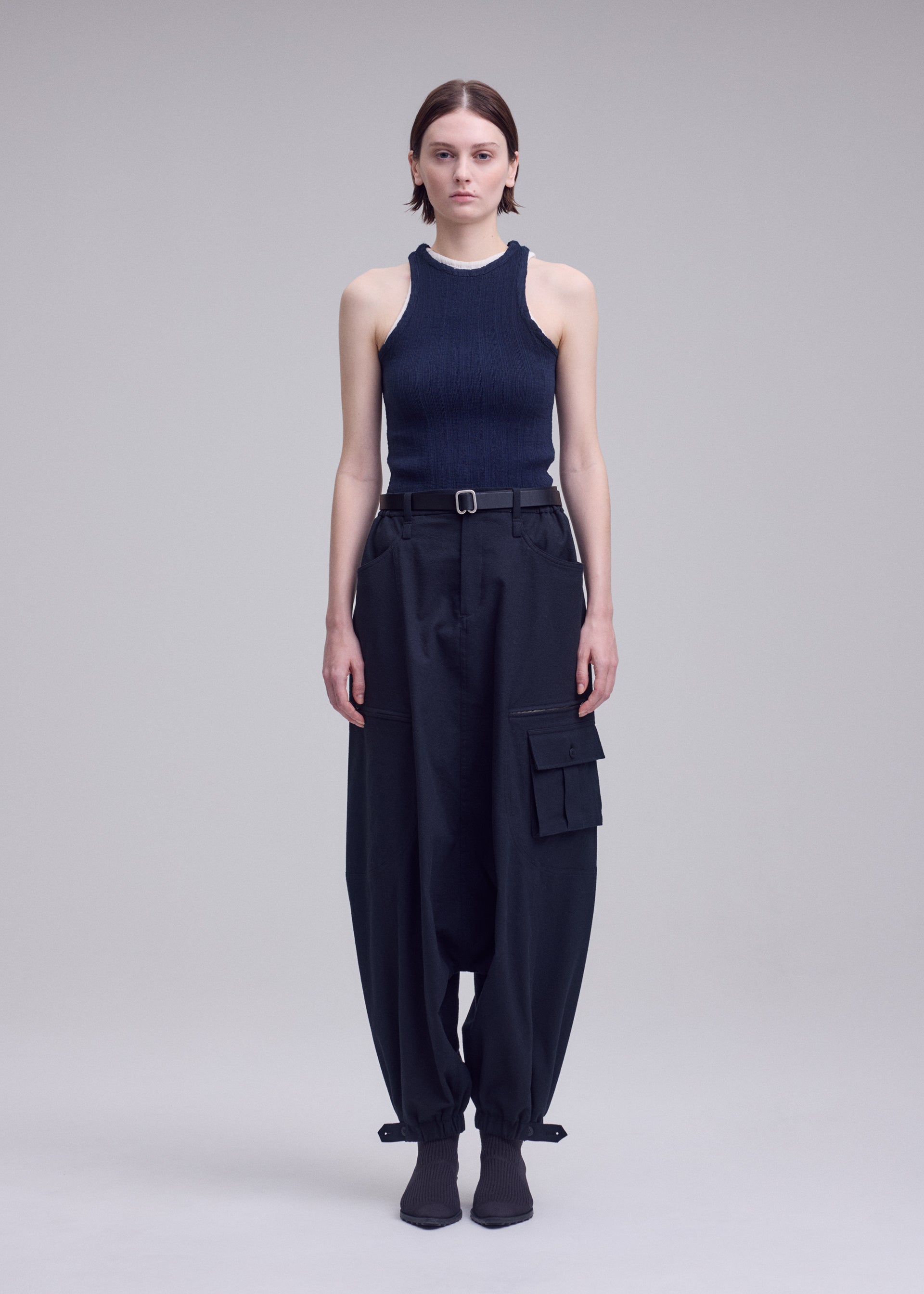 Zoomed view A person with straight brown hair stands against a plain background, wearing a sleeveless black ribbed top, ISSEY MIYAKE GRAINY PANTS featuring jacquard weaving and large pockets, styled with a belt and heeled boots.