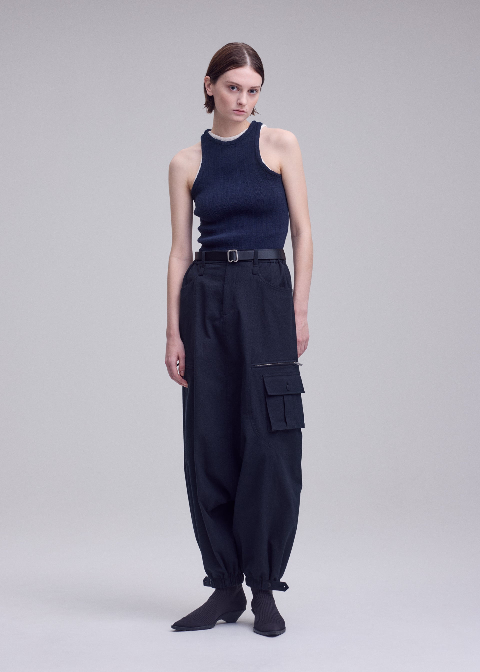 Zoomed view A person stands against a plain background wearing ISSEY MIYAKE’s GRAINY PANTS—high-waisted stretch cotton cargo pants with pockets—paired with a sleeveless black top and black pointed-toe shoes for a modern, minimalist look.