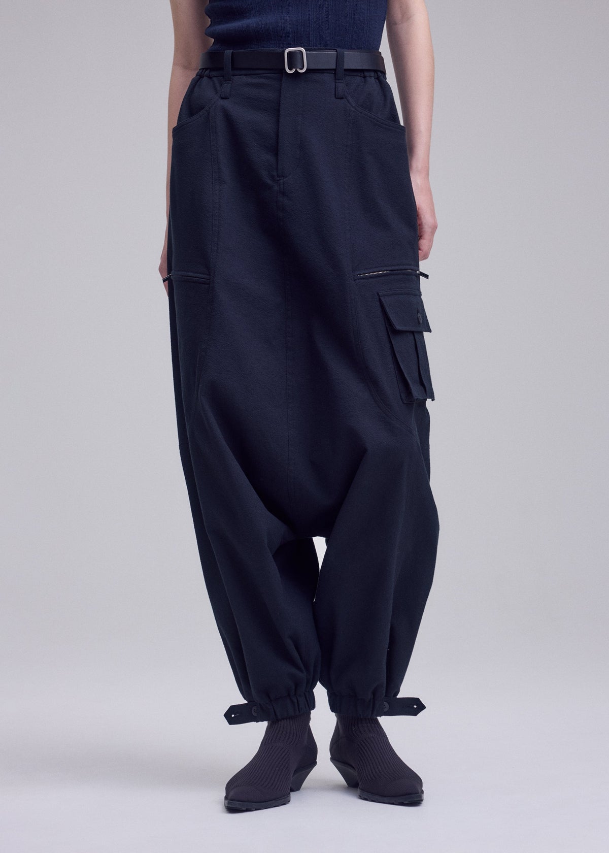 A person wears ISSEY MIYAKE GRAINY PANTS—dark, loose-fitting stretch cotton cargos with zippered pockets, buckled ankle cuffs, and a waist belt—paired with dark socks and a tucked-in dark top. view 8 of 8