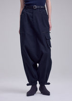 A person wears ISSEY MIYAKE GRAINY PANTS—dark, loose-fitting stretch cotton cargos with zippered pockets, buckled ankle cuffs, and a waist belt—paired with dark socks and a tucked-in dark top.