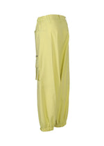 The GRAINY PANTS by ISSEY MIYAKE are light yellow stretch cotton sarouel pants with an elastic waistband, ankle cuffs, side pockets, and a zippered flap pocket on the left leg, shown from the side against a white background.