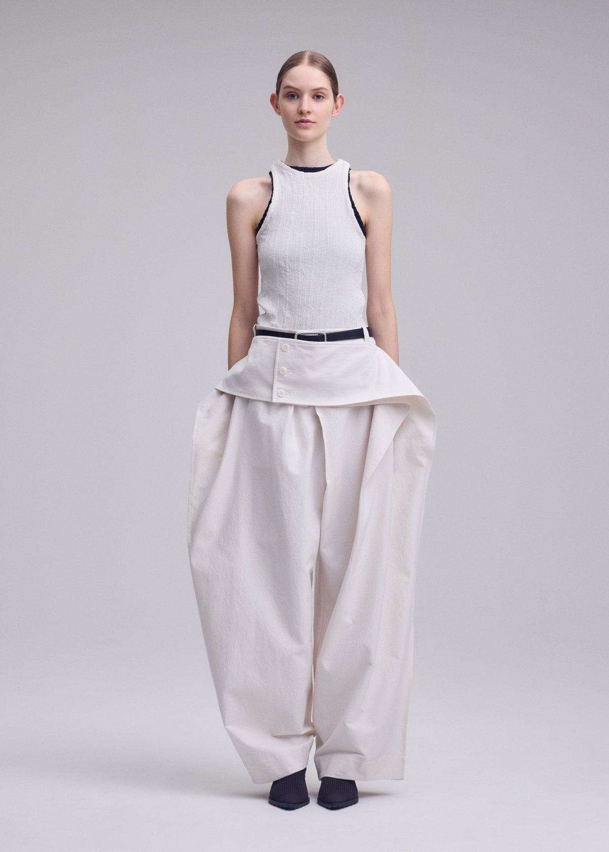 A woman faces forward in ISSEY MIYAKE's GRAINY PANTS—wide, high-waisted jacquard trousers with a layered button detail and connected waistband—worn with a sleeveless ribbed white top and dark shoes, against a plain background. view 5 of 9