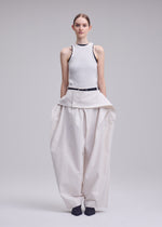A woman faces forward in ISSEY MIYAKE's GRAINY PANTS—wide, high-waisted jacquard trousers with a layered button detail and connected waistband—worn with a sleeveless ribbed white top and dark shoes, against a plain background.