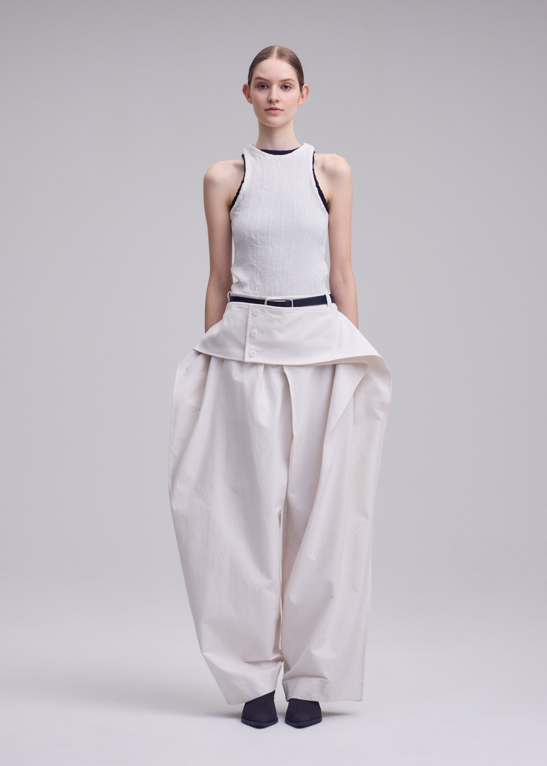 Zoomed view A woman faces forward in ISSEY MIYAKE's GRAINY PANTS—wide, high-waisted jacquard trousers with a layered button detail and connected waistband—worn with a sleeveless ribbed white top and dark shoes, against a plain background.