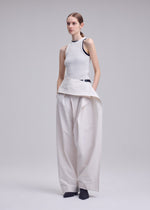 A person stands against a plain light gray background, hands in pockets, wearing ISSEY MIYAKE's GRAINY PANTS—wide-legged, high-waisted jacquard pants with an asymmetrical detail—paired with a sleeveless white top and dark shoes.