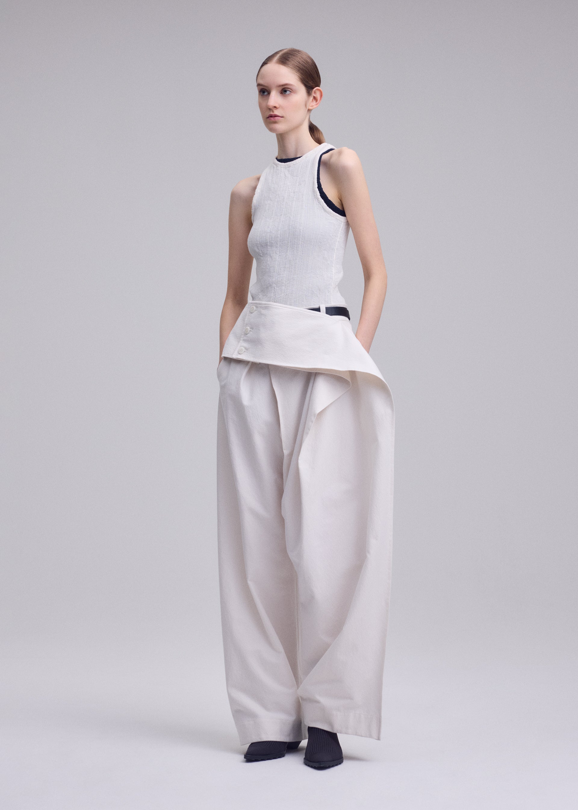 Zoomed view A person stands against a plain light gray background, hands in pockets, wearing ISSEY MIYAKE's GRAINY PANTS—wide-legged, high-waisted jacquard pants with an asymmetrical detail—paired with a sleeveless white top and dark shoes.