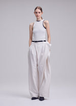 A woman poses against a plain background in ISSEY MIYAKE GRAINY PANTS, wearing a sleeveless white ribbed top. The high-waisted pants feature a connected waistband and black belt. She completes the look with black shoes and hands in her pockets.