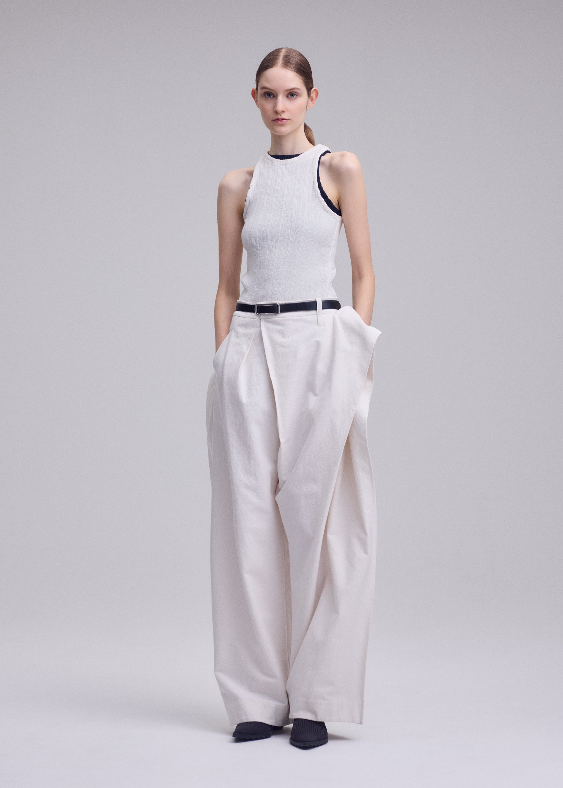 Zoomed view A woman poses against a plain background in ISSEY MIYAKE GRAINY PANTS, wearing a sleeveless white ribbed top. The high-waisted pants feature a connected waistband and black belt. She completes the look with black shoes and hands in her pockets.
