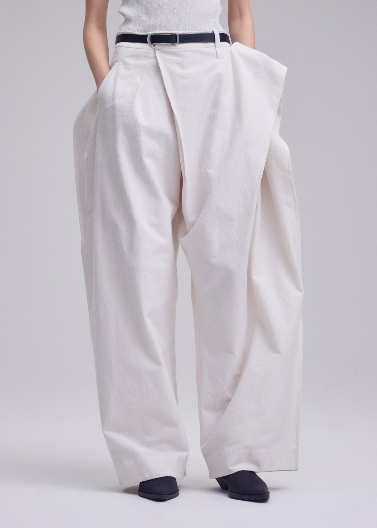 A person wears ISSEY MIYAKE’s GRAINY PANTS—oversized, high-waisted white jacquard trousers with a sculptural fit, connected waistband, and pleats—styled with a black belt and black pointed shoes against a plain light background. view 8 of 9