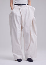 A person wears ISSEY MIYAKE’s GRAINY PANTS—oversized, high-waisted white jacquard trousers with a sculptural fit, connected waistband, and pleats—styled with a black belt and black pointed shoes against a plain light background.
