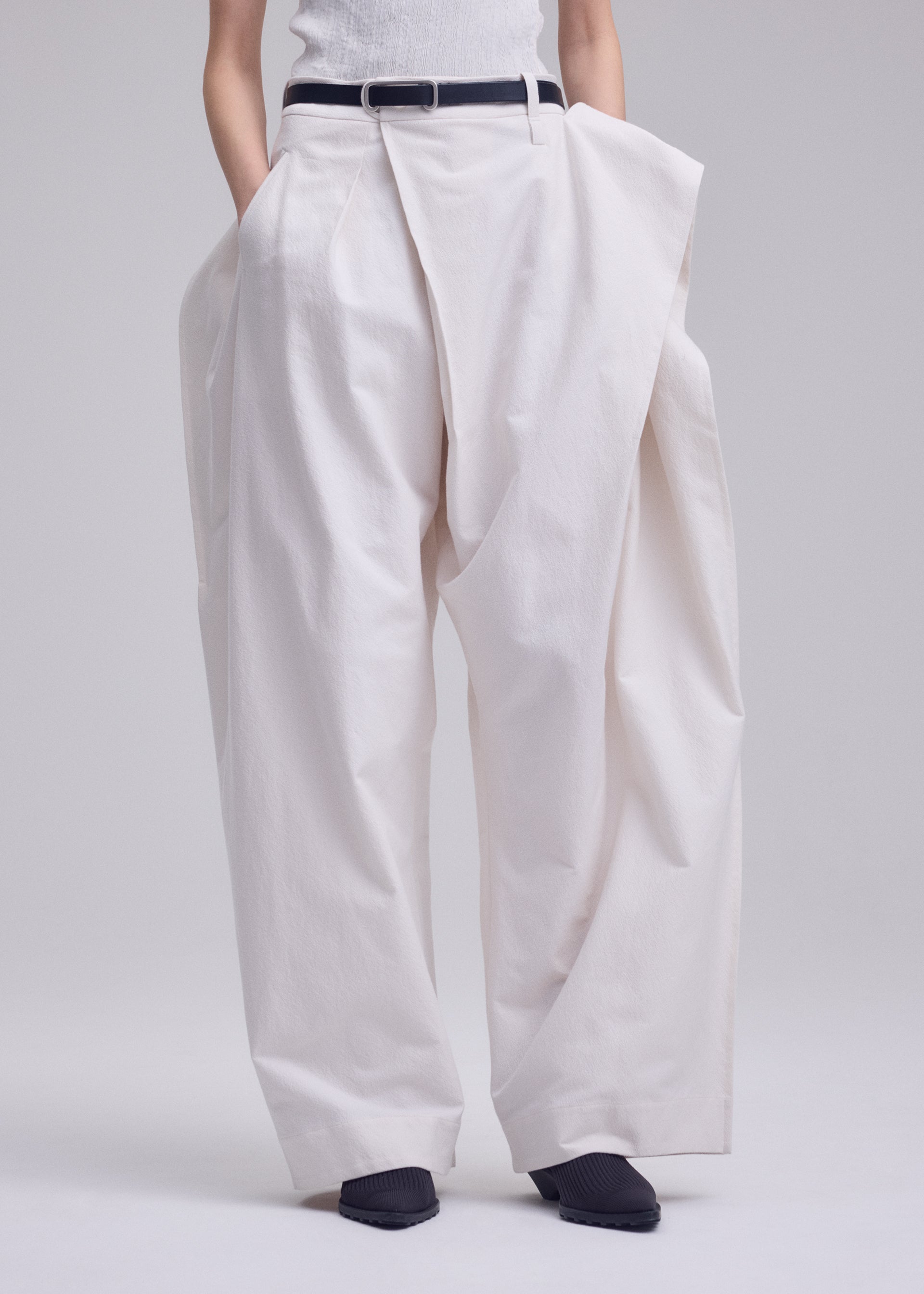 Zoomed view A person wears ISSEY MIYAKE’s GRAINY PANTS—oversized, high-waisted white jacquard trousers with a sculptural fit, connected waistband, and pleats—styled with a black belt and black pointed shoes against a plain light background.