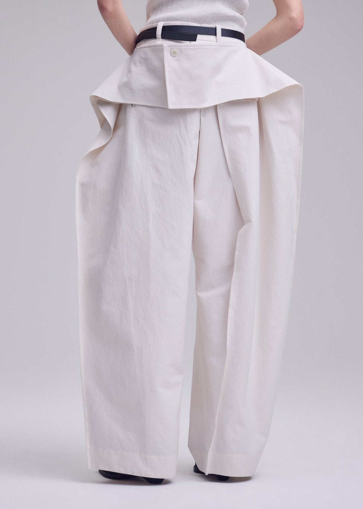Wearing ISSEY MIYAKE GRAINY PANTS—high-waisted, wide-leg jacquard pants with exaggerated volume and a skirt-like flap at the waistband—paired with a black belt; their arms are behind their back. view 9 of 9