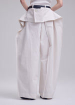 Wearing ISSEY MIYAKE GRAINY PANTS—high-waisted, wide-leg jacquard pants with exaggerated volume and a skirt-like flap at the waistband—paired with a black belt; their arms are behind their back.