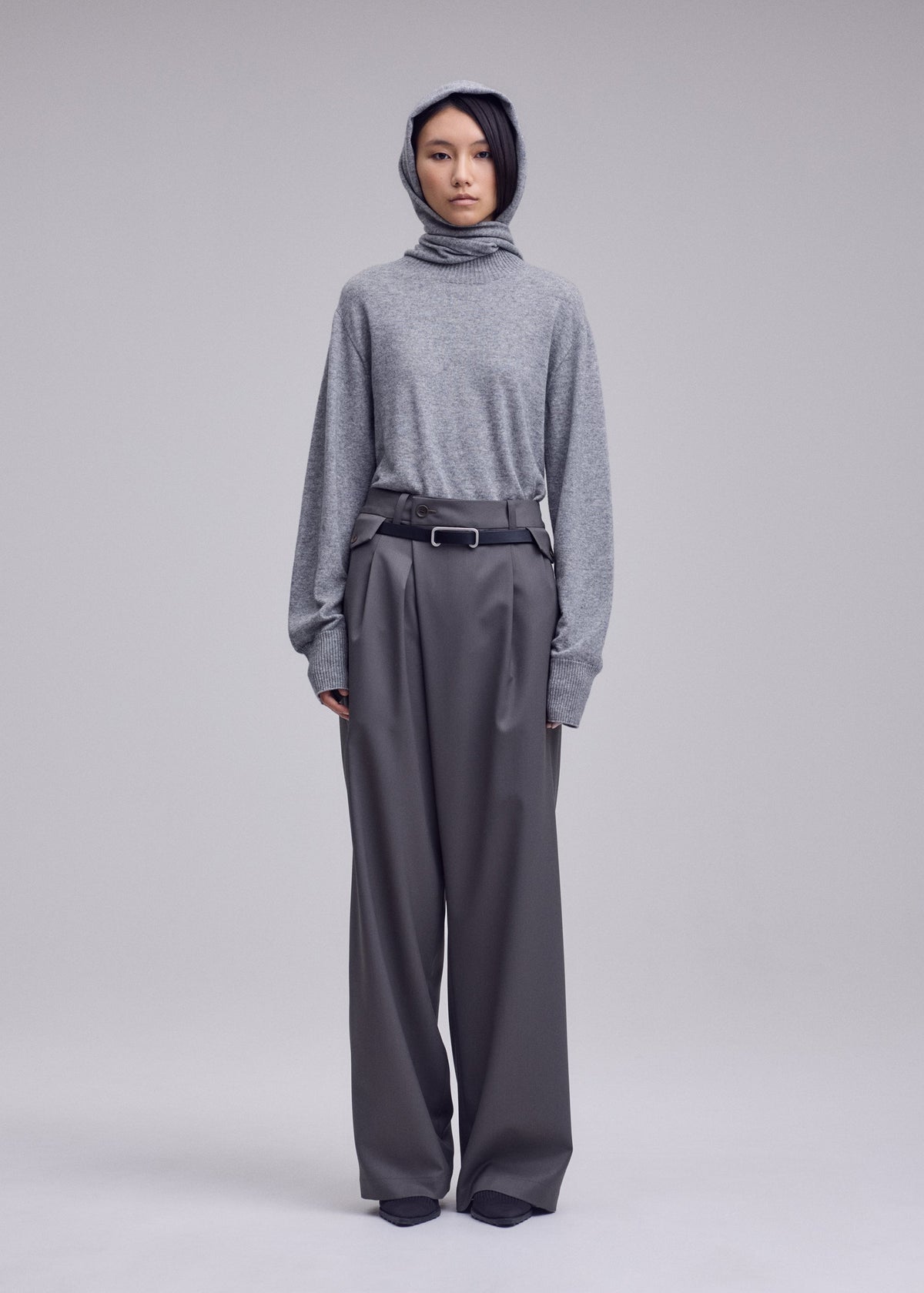 A person stands facing forward against a light gray studio background, wearing a gray hooded sweater paired with ISSEY MIYAKE LUSTROUS WOOL PANTS—loose, high-waisted, wide gabardine trousers with a belt. Crafted from a wrinkle-resistant wool blend that provides both structure and ease, these versatile pants are also hand-washable for effortless care. view 6 of 8