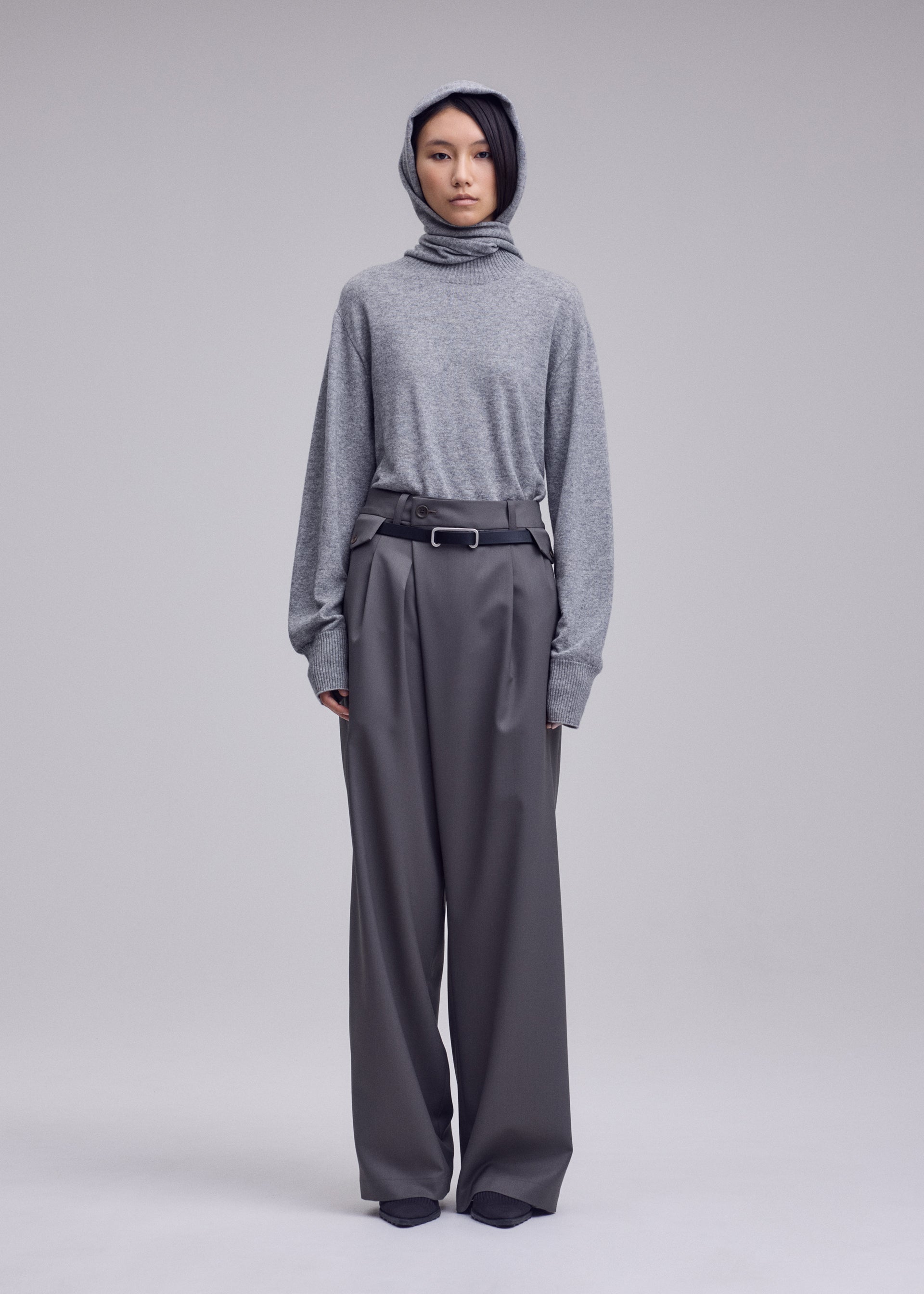 Zoomed view A person stands facing forward against a light gray studio background, wearing a gray hooded sweater paired with ISSEY MIYAKE LUSTROUS WOOL PANTS—loose, high-waisted, wide gabardine trousers with a belt. Crafted from a wrinkle-resistant wool blend that provides both structure and ease, these versatile pants are also hand-washable for effortless care.