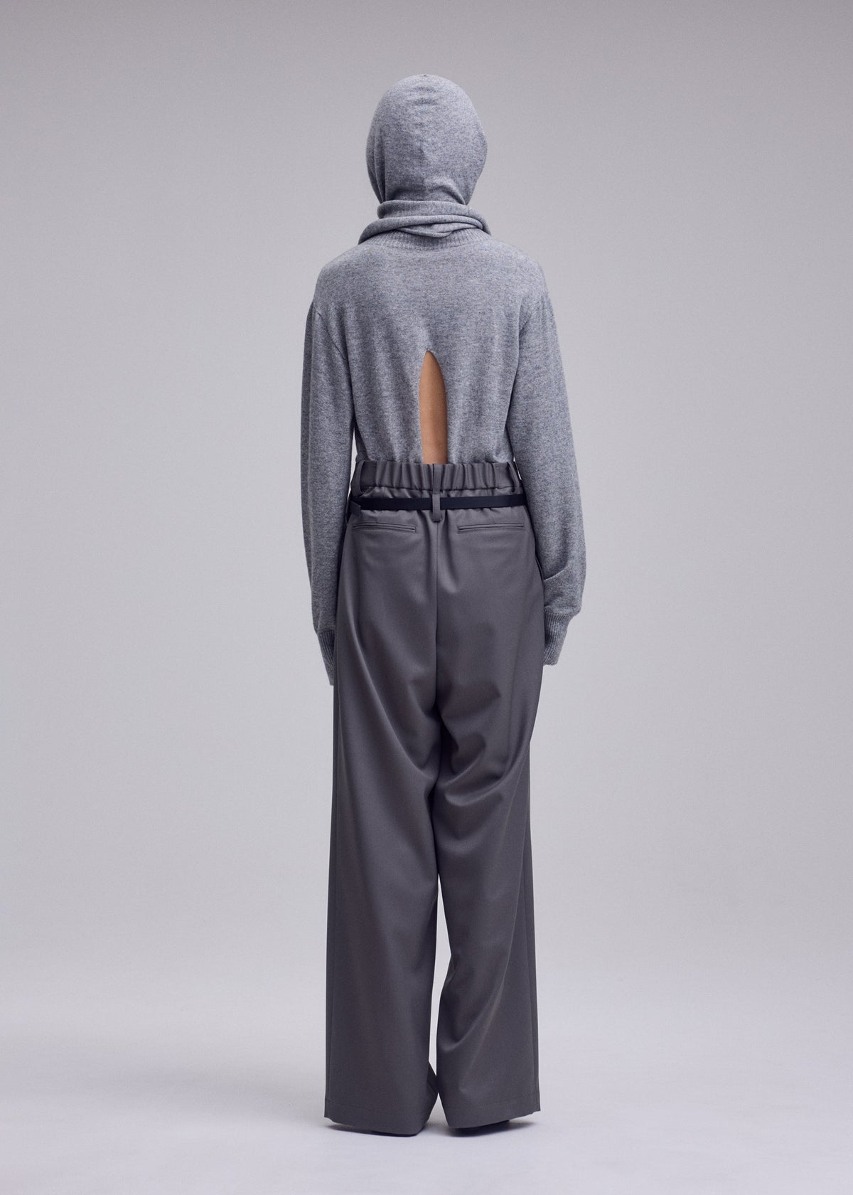 A person faces away, wearing a gray hooded sweater with a back slit and loose, high-waisted LUSTROUS WOOL PANTS by ISSEY MIYAKE, crafted from a wrinkle-resistant wool blend. The background is plain and light-colored. view 7 of 8