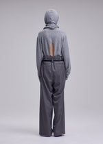 A person faces away, wearing a gray hooded sweater with a back slit and loose, high-waisted LUSTROUS WOOL PANTS by ISSEY MIYAKE, crafted from a wrinkle-resistant wool blend. The background is plain and light-colored.
