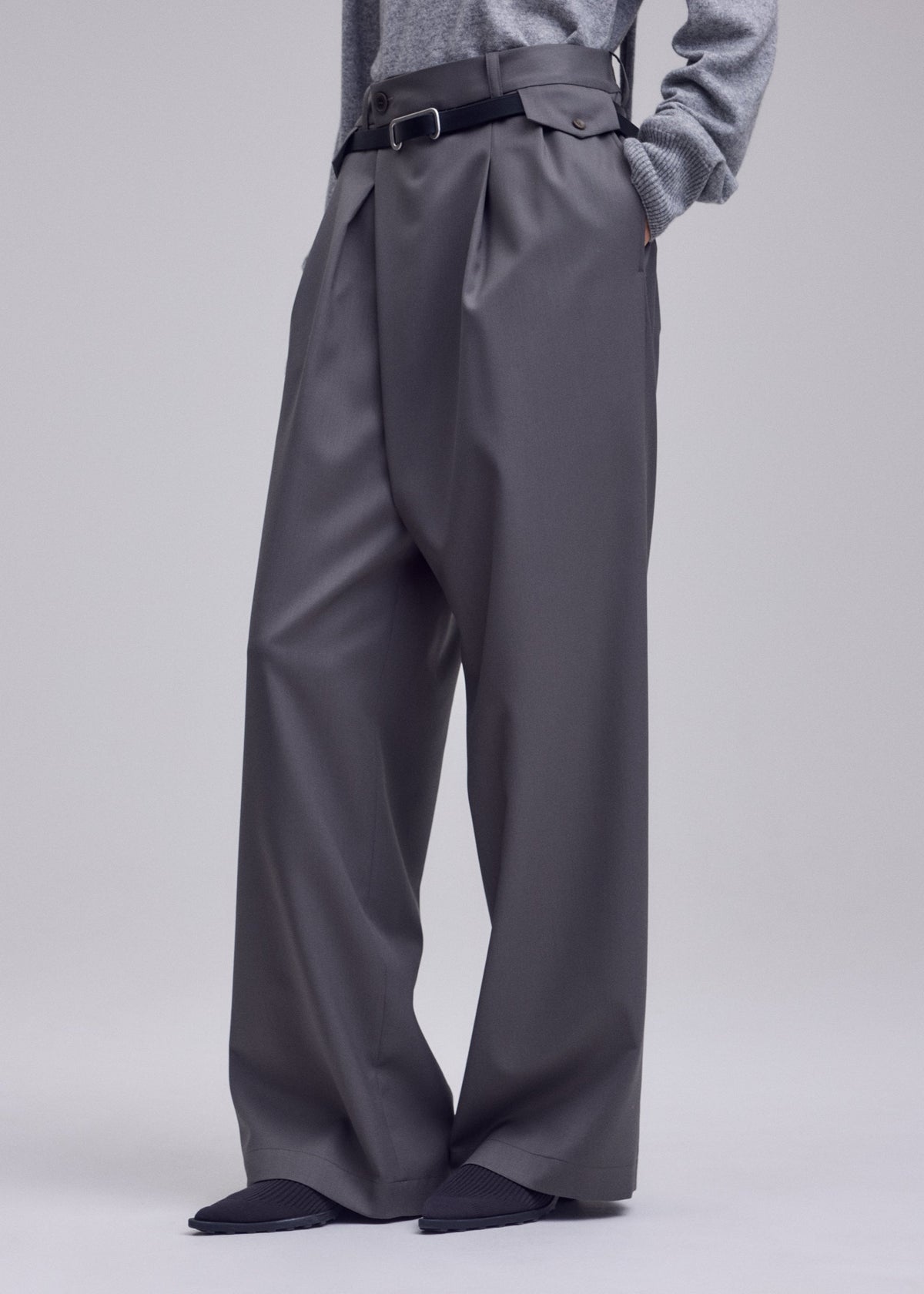 A person wearing ISSEY MIYAKE LUSTROUS WOOL PANTS—wide, wrinkle-resistant gabardine trousers—styled with a black belt, gray top, and pointed black shoes, stands hands-in-pockets against a plain background. view 8 of 8