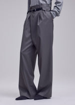 A person wearing ISSEY MIYAKE LUSTROUS WOOL PANTS—wide, wrinkle-resistant gabardine trousers—styled with a black belt, gray top, and pointed black shoes, stands hands-in-pockets against a plain background.