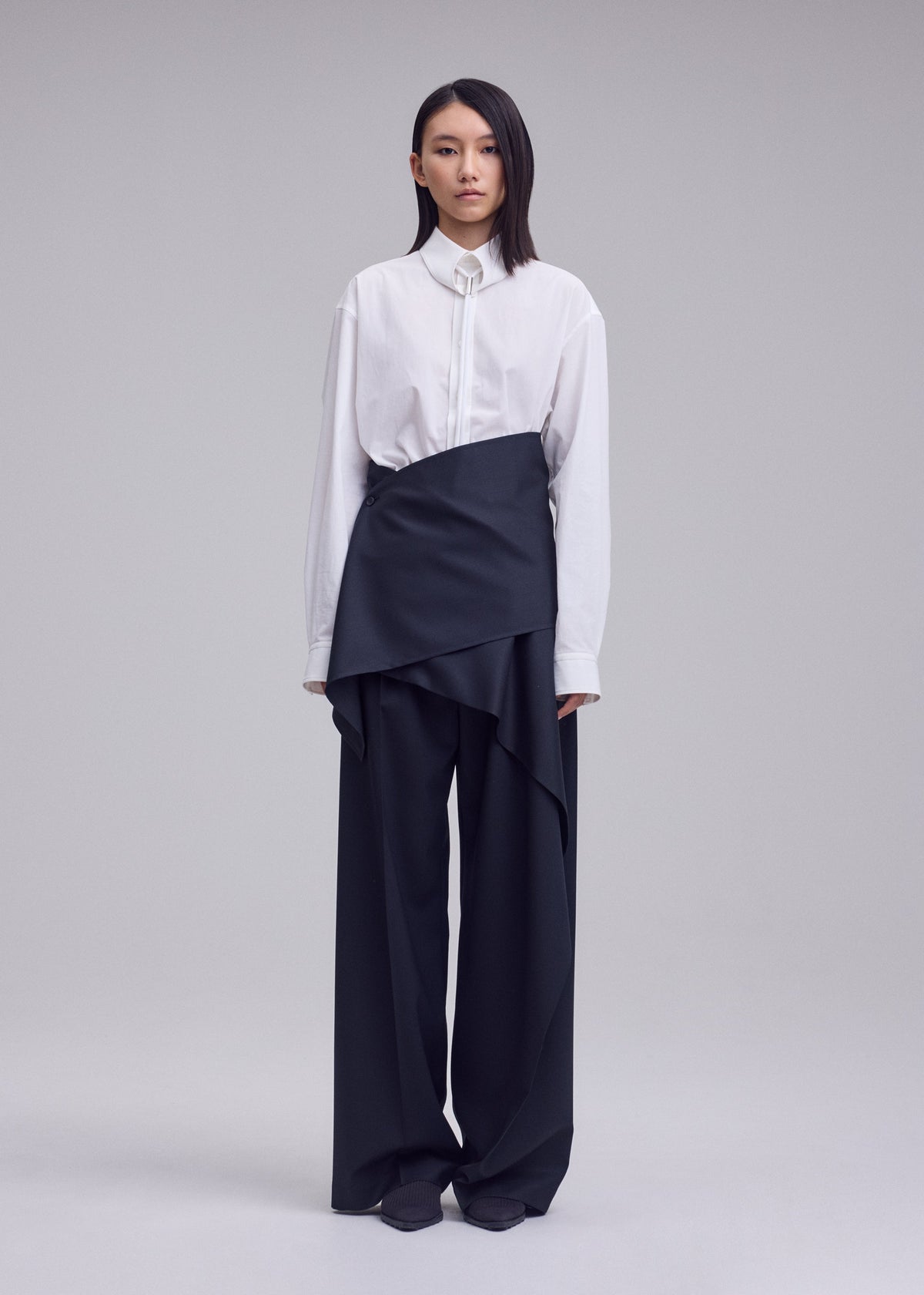 A person stands against a plain background, wearing a white button-up shirt paired with the ISSEY MIYAKE LUSTROUS WOOL PANTS—loose, wide-leg black gabardine trousers featuring an asymmetrical layered wrap design. The wrinkle-resistant wool blend fabric offers structure and effortless style to the look. Their straight, shoulder-length dark hair frames the ensemble. view 6 of 9