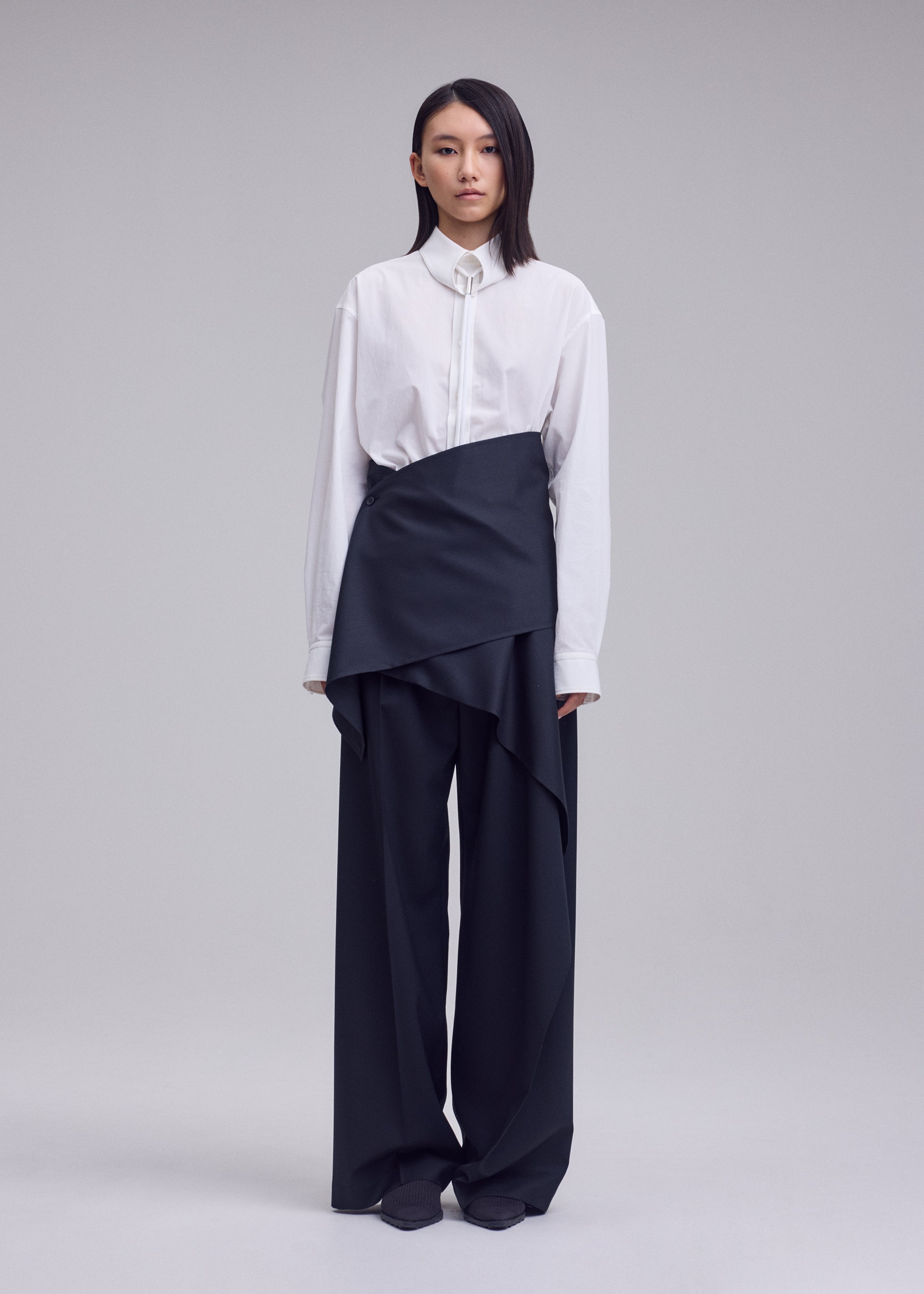Zoomed view A person stands against a plain background, wearing a white button-up shirt paired with the ISSEY MIYAKE LUSTROUS WOOL PANTS—loose, wide-leg black gabardine trousers featuring an asymmetrical layered wrap design. The wrinkle-resistant wool blend fabric offers structure and effortless style to the look. Their straight, shoulder-length dark hair frames the ensemble.