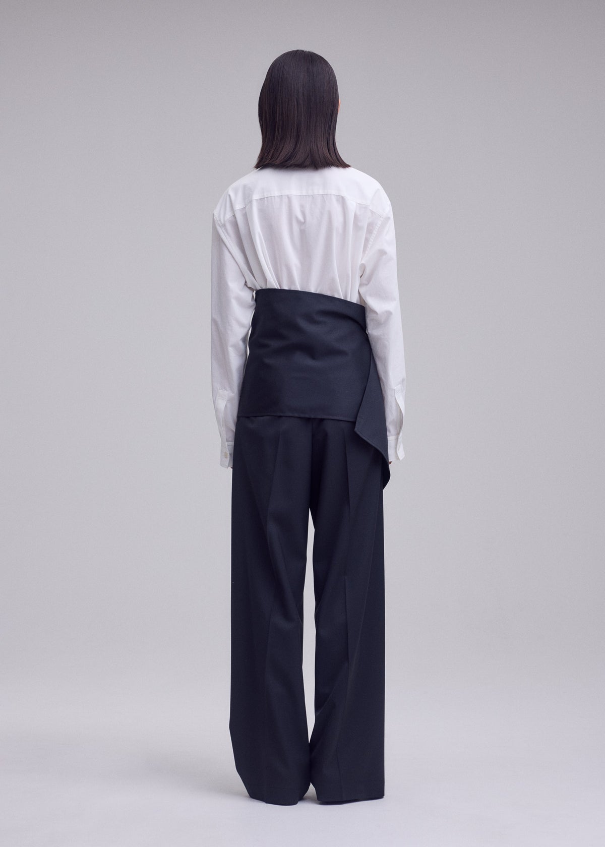 A person stands facing away, wearing a white button-up shirt and ISSEY MIYAKE’s LUSTROUS WOOL PANTS—wide-leg, black trousers crafted from a wrinkle-resistant wool blend that offers a polished look. A black sash or wrap is tied over their shirt at the waist, standing out against the plain, light-colored background. view 7 of 9