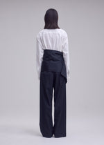 A person stands facing away, wearing a white button-up shirt and ISSEY MIYAKE’s LUSTROUS WOOL PANTS—wide-leg, black trousers crafted from a wrinkle-resistant wool blend that offers a polished look. A black sash or wrap is tied over their shirt at the waist, standing out against the plain, light-colored background.