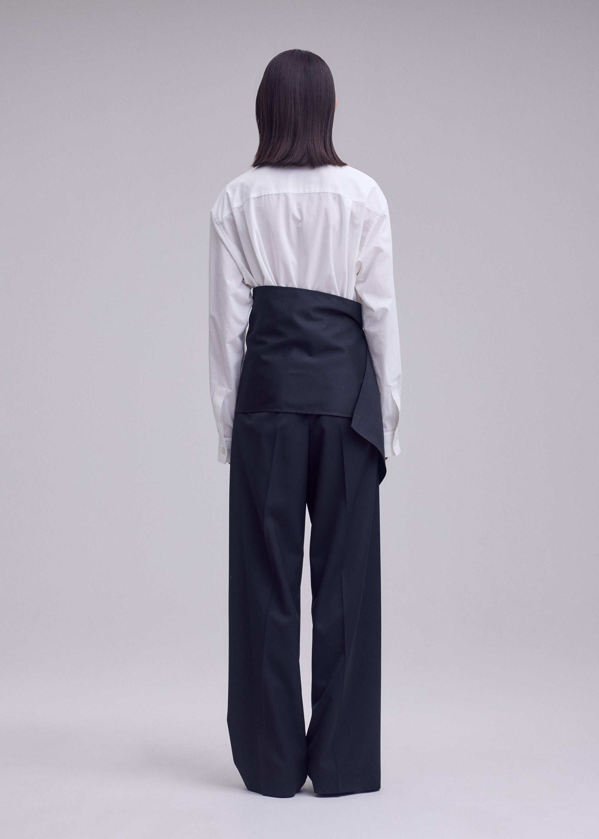 Zoomed view A person stands facing away, wearing a white button-up shirt and ISSEY MIYAKE’s LUSTROUS WOOL PANTS—wide-leg, black trousers crafted from a wrinkle-resistant wool blend that offers a polished look. A black sash or wrap is tied over their shirt at the waist, standing out against the plain, light-colored background.