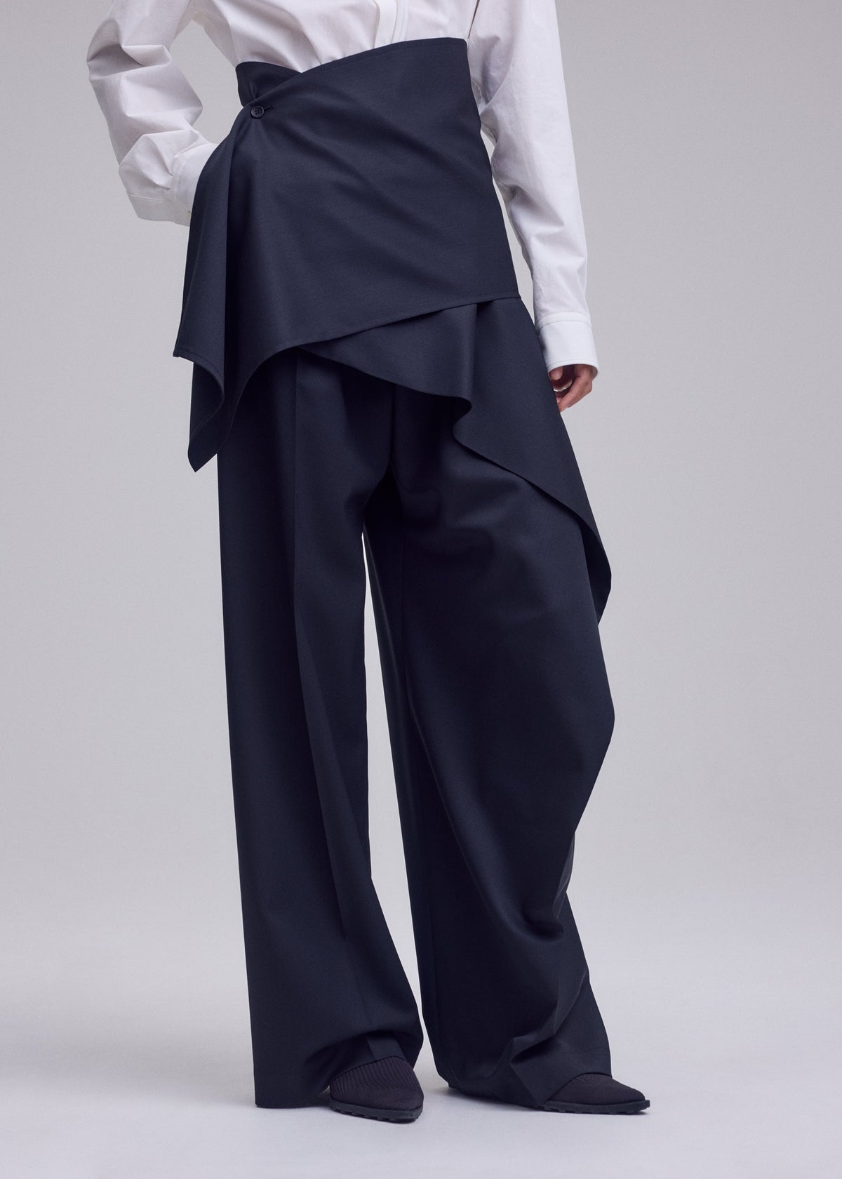 A person wearing ISSEY MIYAKE LUSTROUS WOOL PANTS featuring a wide-leg silhouette with a layered, asymmetrical overskirt, paired with a white dress shirt. Only the body from shoulders down is visible. view 8 of 9