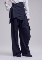 A person wearing ISSEY MIYAKE LUSTROUS WOOL PANTS featuring a wide-leg silhouette with a layered, asymmetrical overskirt, paired with a white dress shirt. Only the body from shoulders down is visible.