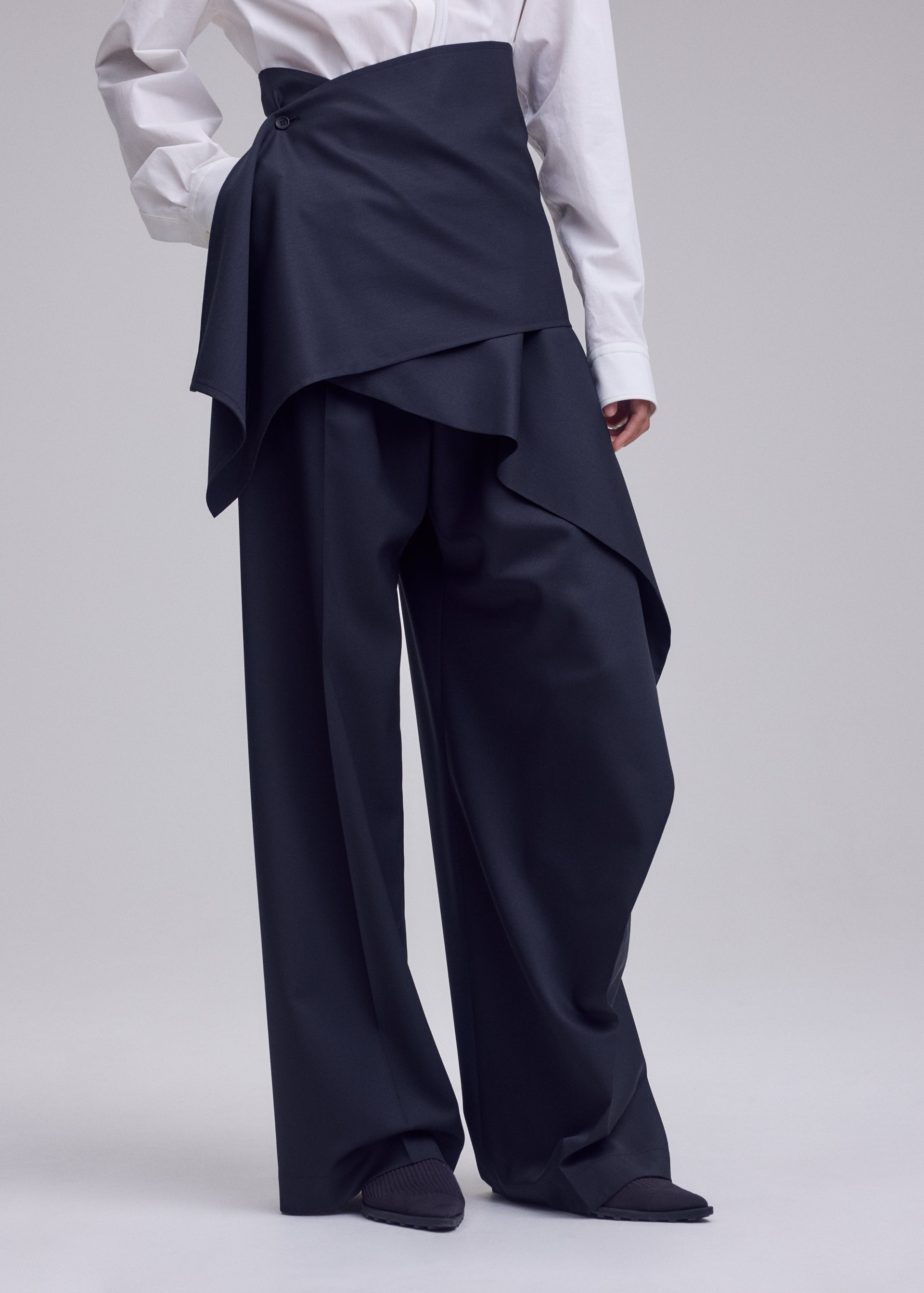 Zoomed view A person wearing ISSEY MIYAKE LUSTROUS WOOL PANTS featuring a wide-leg silhouette with a layered, asymmetrical overskirt, paired with a white dress shirt. Only the body from shoulders down is visible.
