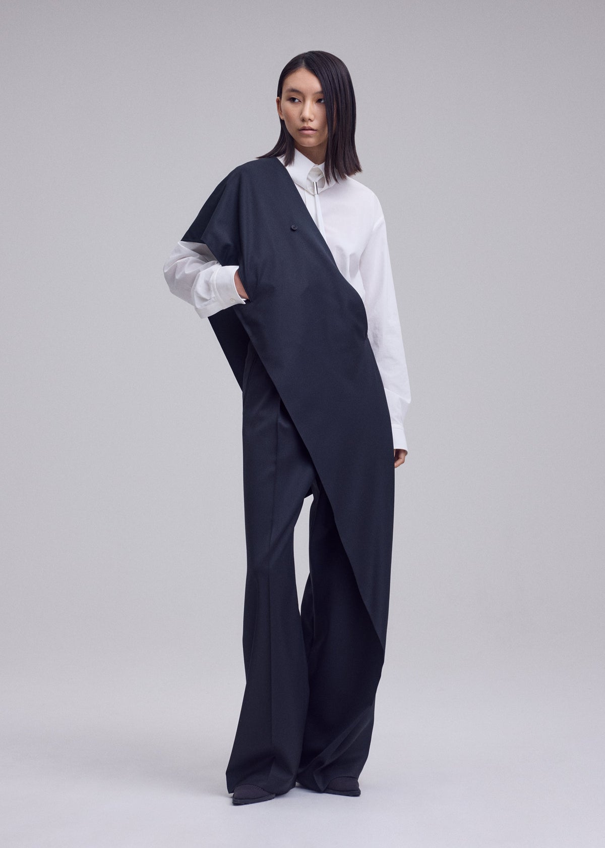A person stands in a studio wearing a white button-up shirt and an oversized, asymmetrical black garment draped across the body, paired with ISSEY MIYAKE LUSTROUS WOOL PANTS. The background is plain and light-colored. view 9 of 9