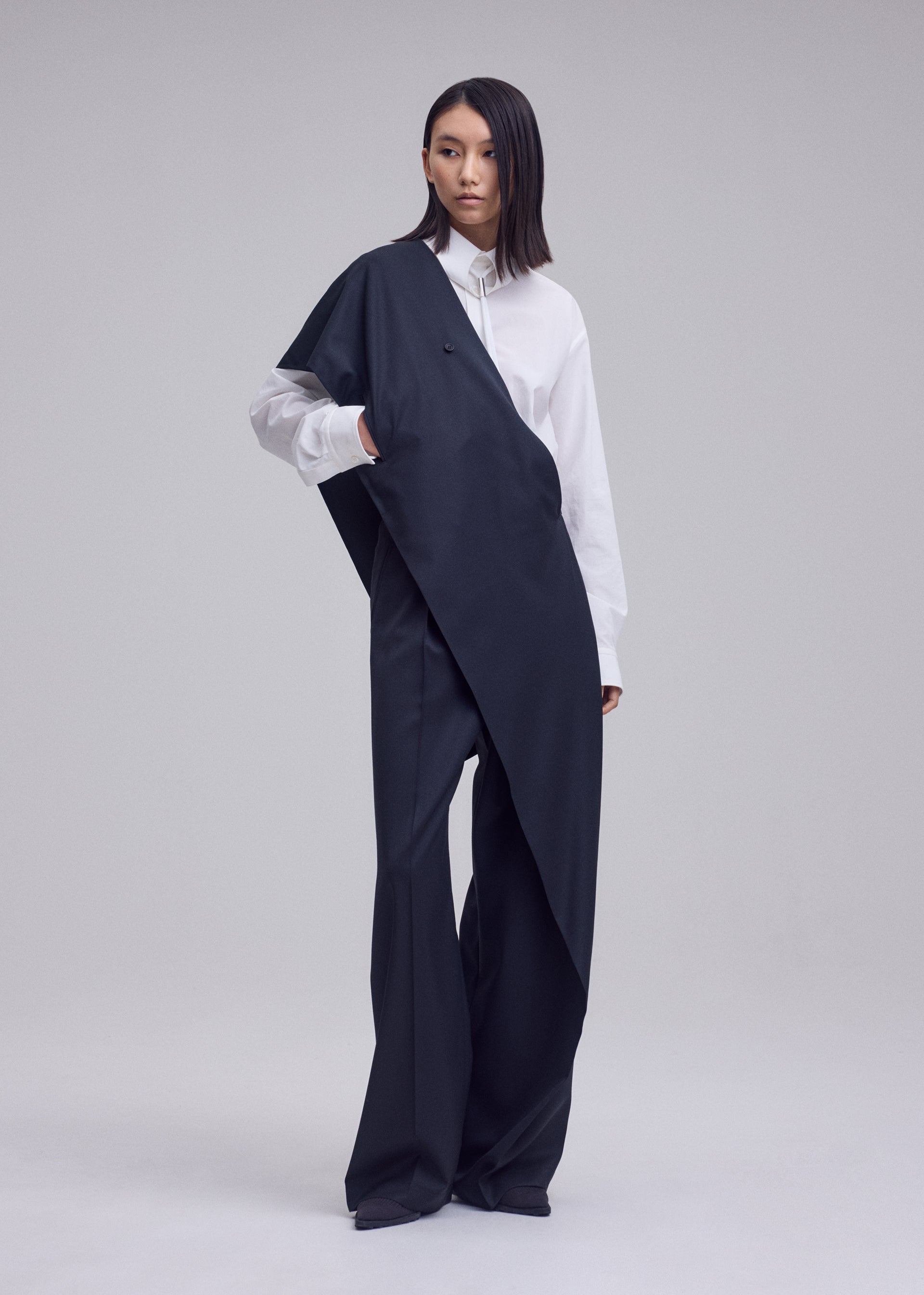Zoomed view A person stands in a studio wearing a white button-up shirt and an oversized, asymmetrical black garment draped across the body, paired with ISSEY MIYAKE LUSTROUS WOOL PANTS. The background is plain and light-colored.