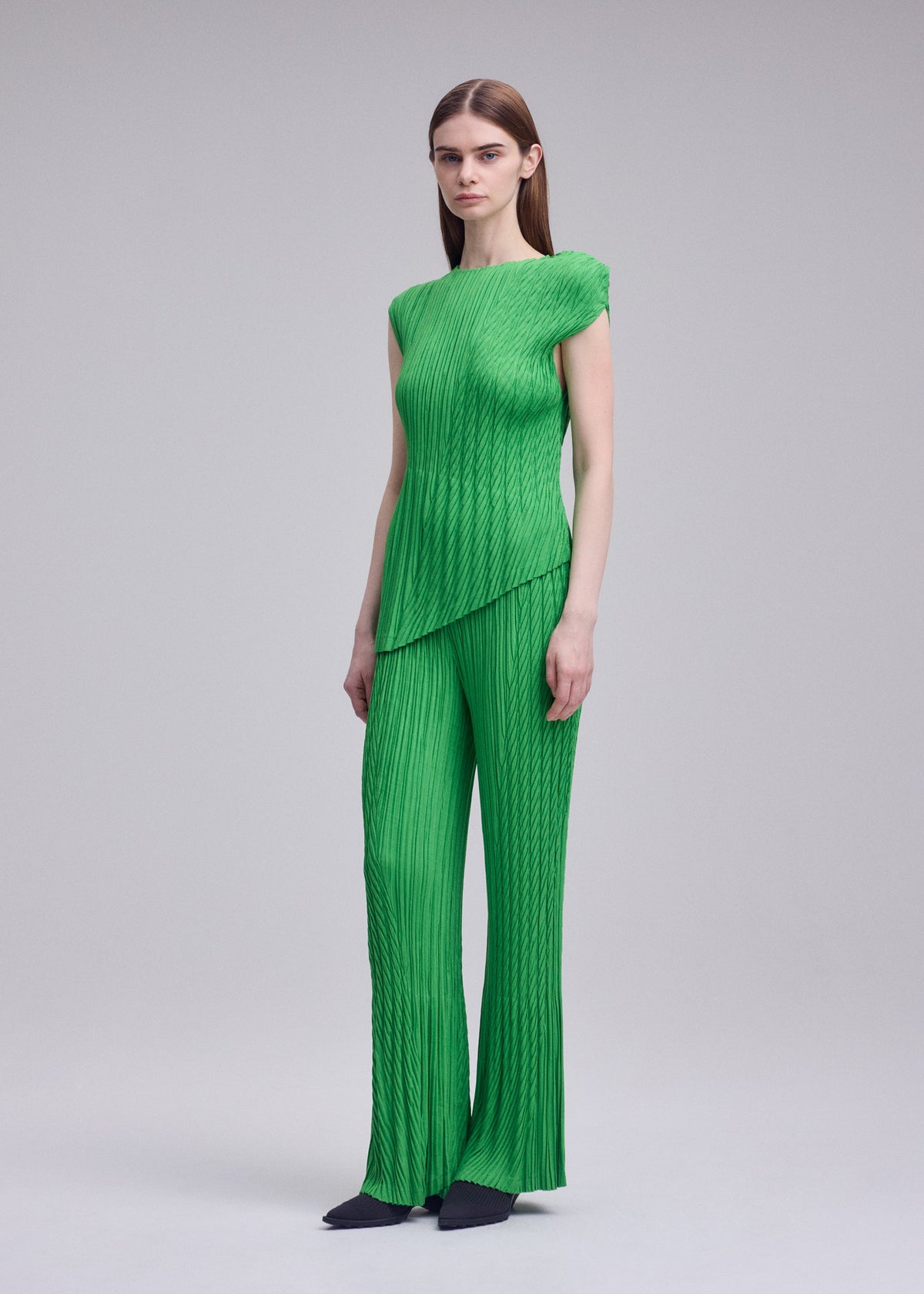 A woman stands in a studio wearing ISSEY MIYAKE’s UNFURLING PLEATS set—a bright green pleated top and flared pants. She has straight brown hair, black shoes, and the background is plain light gray. view 7 of 8