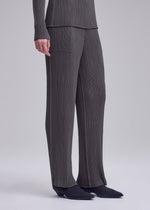 Model showcases ISSEY MIYAKE’s HATCHING PLEATS—gray, textured pleated straight pants with a matching long-sleeve top. Only the body from shoulders down and black-shod feet are shown, against a light backdrop.