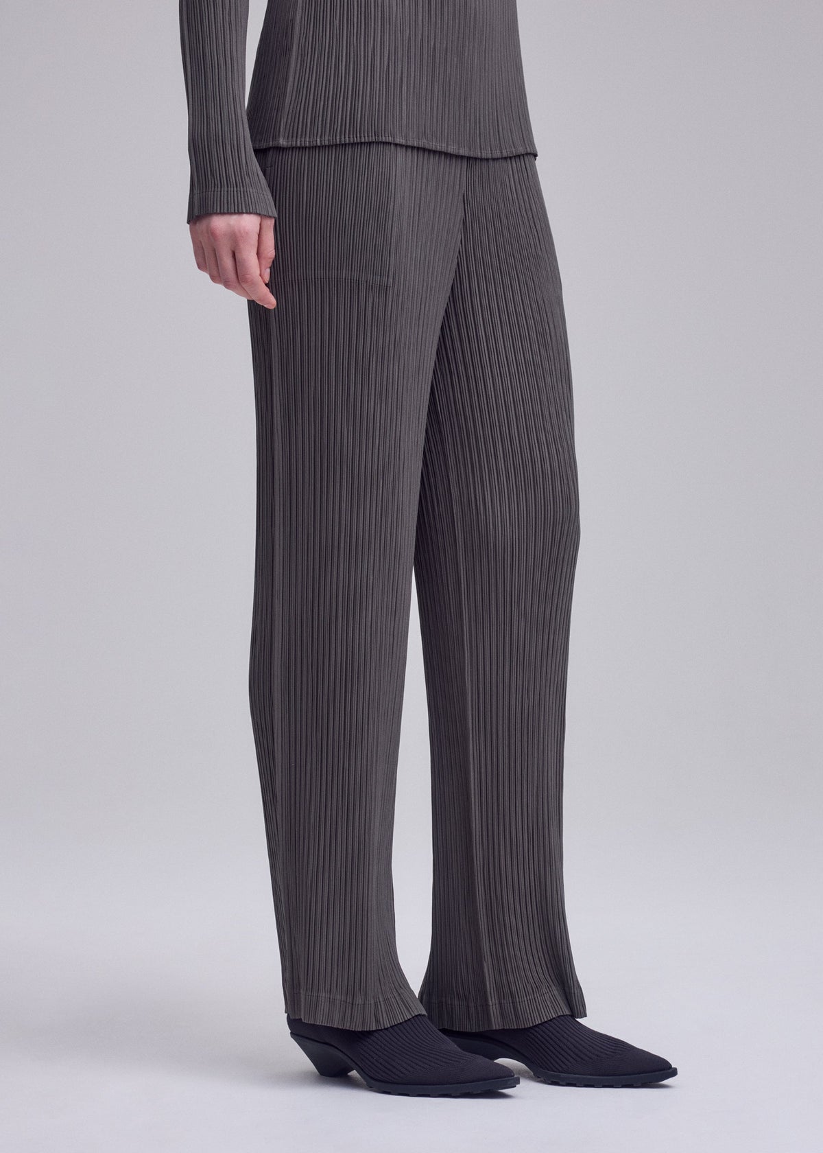 Zoomed view Model showcases ISSEY MIYAKE’s HATCHING PLEATS—gray, textured pleated straight pants with a matching long-sleeve top. Only the body from shoulders down and black-shod feet are shown, against a light backdrop.