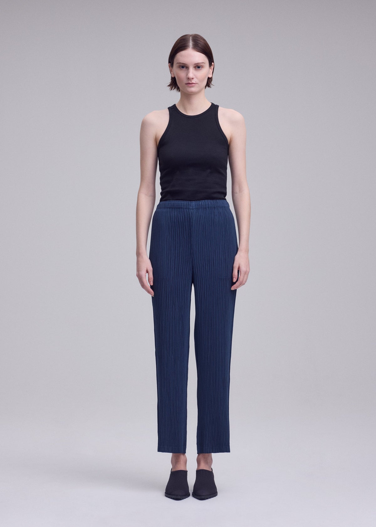 A person stands facing forward against a plain light background, wearing a black sleeveless top, ISSEY MIYAKE HATCHING BOTTOMS—blue pleated straight pants with a subtle hatching finish—and black shoes. view 4 of 6