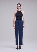 A person stands facing forward against a plain light gray background, wearing a black sleeveless top, black shoes, and the ISSEY MIYAKE HATCHING BOTTOMS—blue pleated straight pants with a hatching appearance.