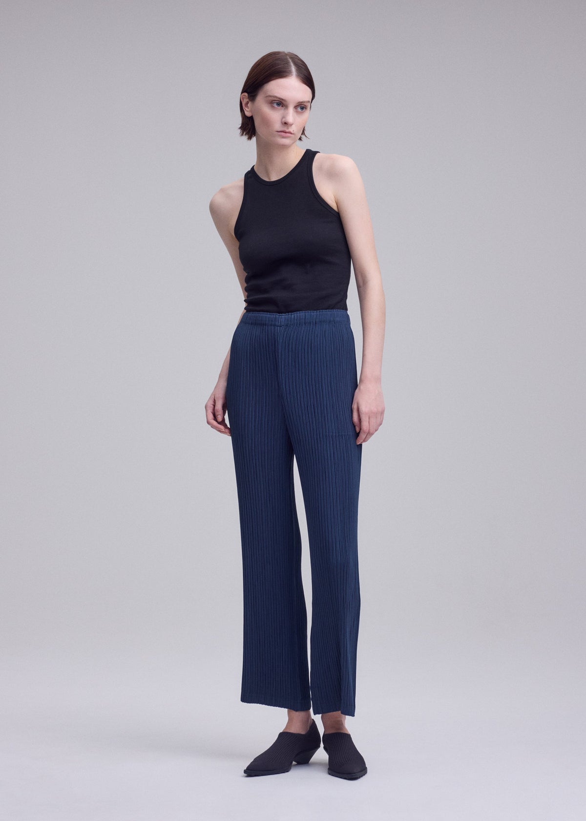 A person with short brown hair wears a black sleeveless top, blue high-waisted textured polyester pants, and black shoes, posing against a plain light gray background. view 5 of 6