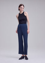 A person with short brown hair wears a black sleeveless top, blue high-waisted textured polyester pants, and black shoes, posing against a plain light gray background.