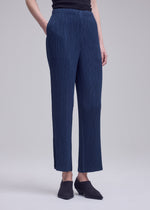 A model showcases the ISSEY MIYAKE HATCHING BOTTOMS—navy blue pleated straight pants crafted from textured polyester—standing against a plain gray background. Only the lower half of the body is visible, with one hand placed in a pocket and black shoes completing the look.