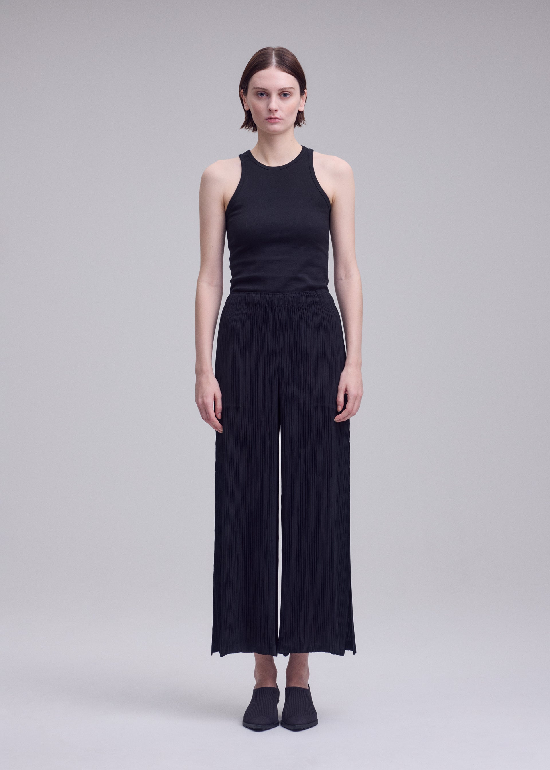 Zoomed view A person stands facing forward against a plain light gray background, wearing black HATCHING BOTTOMS with a textured pleated wide-leg design by ISSEY MIYAKE, paired with a black sleeveless top and black shoes.