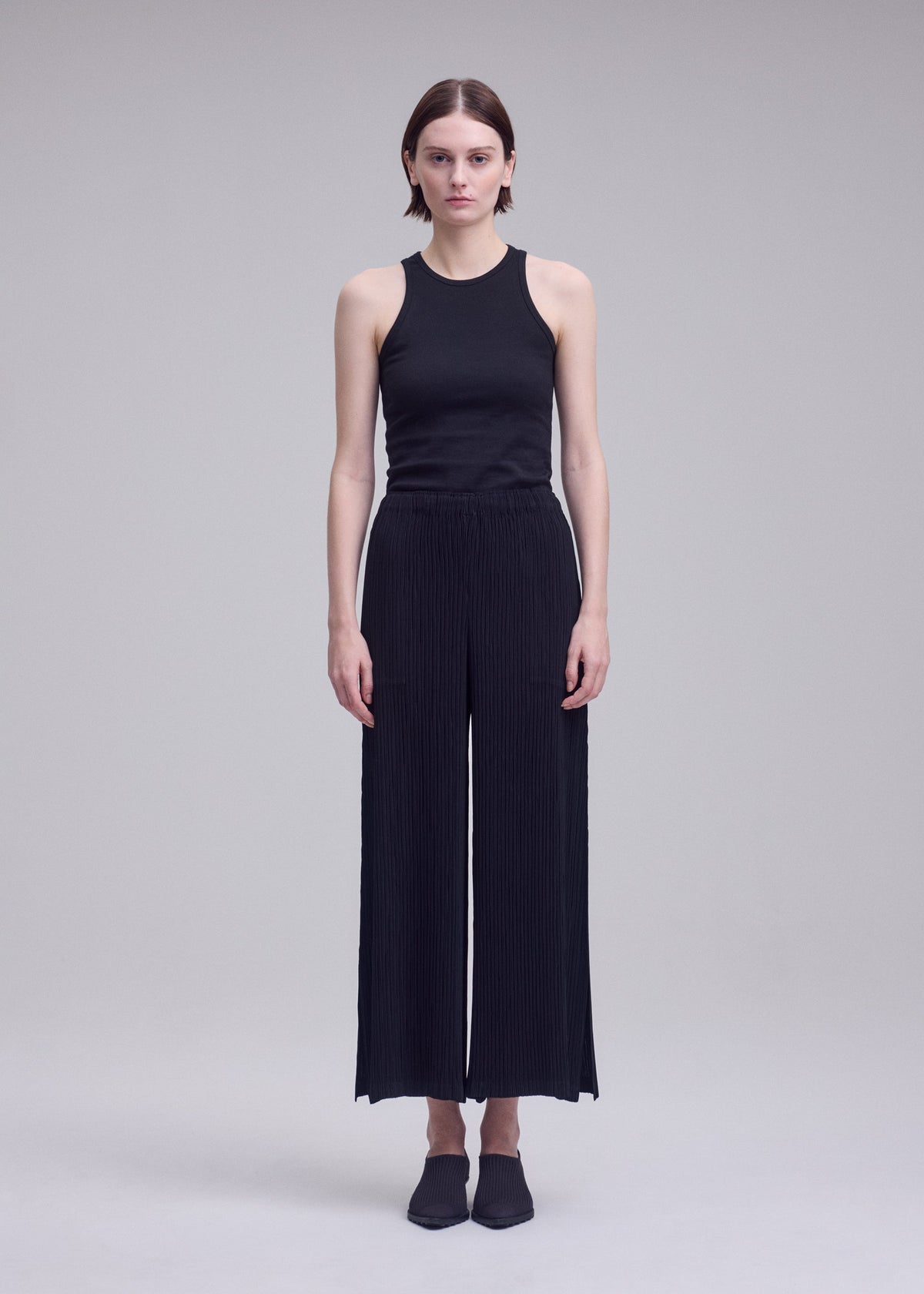 A person stands facing forward against a plain light gray background, wearing HATCHING BOTTOMS by ISSEY MIYAKE—a pair of pleated wide pants—along with a sleeveless black top and black shoes. view 4 of 6