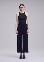 A person stands facing forward against a plain light gray background, wearing HATCHING BOTTOMS by ISSEY MIYAKE—a pair of pleated wide pants—along with a sleeveless black top and black shoes.