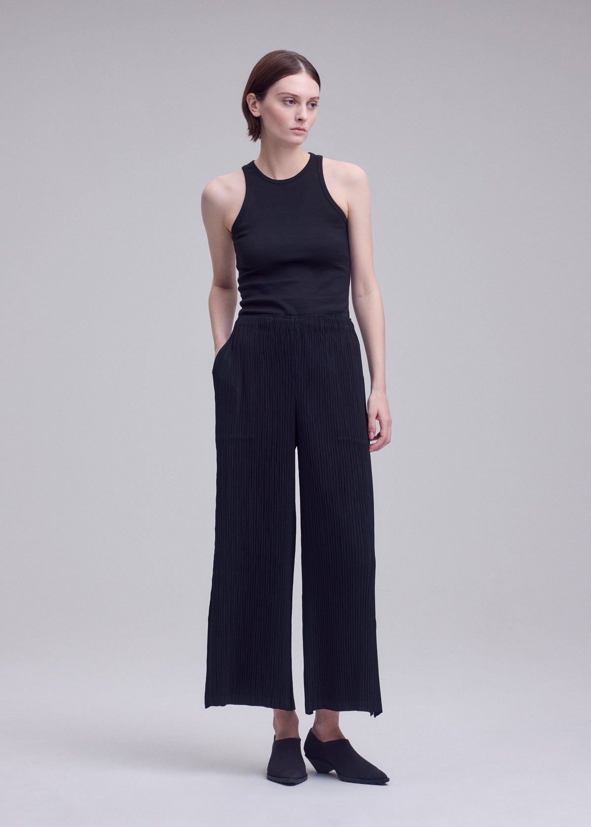 A person stands against a gray background, wearing a sleeveless black top and ISSEY MIYAKE HATCHING BOTTOMS—pleated wide pants in black textured polyester—paired with black shoes. They have short brown hair, hands in their pockets, and are looking slightly to the right. view 5 of 6