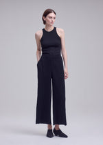 A person stands against a gray background, wearing a sleeveless black top and ISSEY MIYAKE HATCHING BOTTOMS—pleated wide pants in black textured polyester—paired with black shoes. They have short brown hair, hands in their pockets, and are looking slightly to the right.