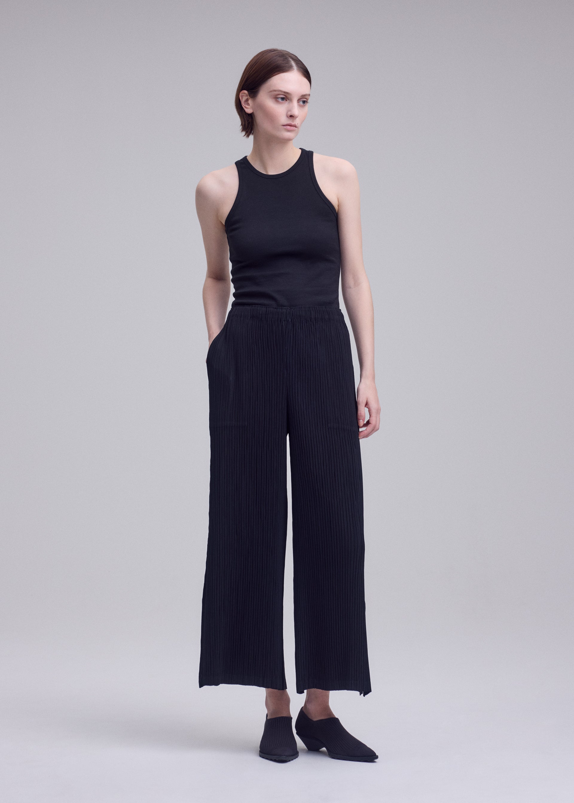 Zoomed view A person stands against a plain background, wearing a sleeveless black top paired with ISSEY MIYAKE’s HATCHING BOTTOMS—pleated wide-leg pants with pockets crafted from polyester. Their short brown hair is styled simply as they gaze slightly to the side, while the ensemble’s subtle textured finish lends visual interest to the minimalist look. Black slip-on shoes complete the outfit.