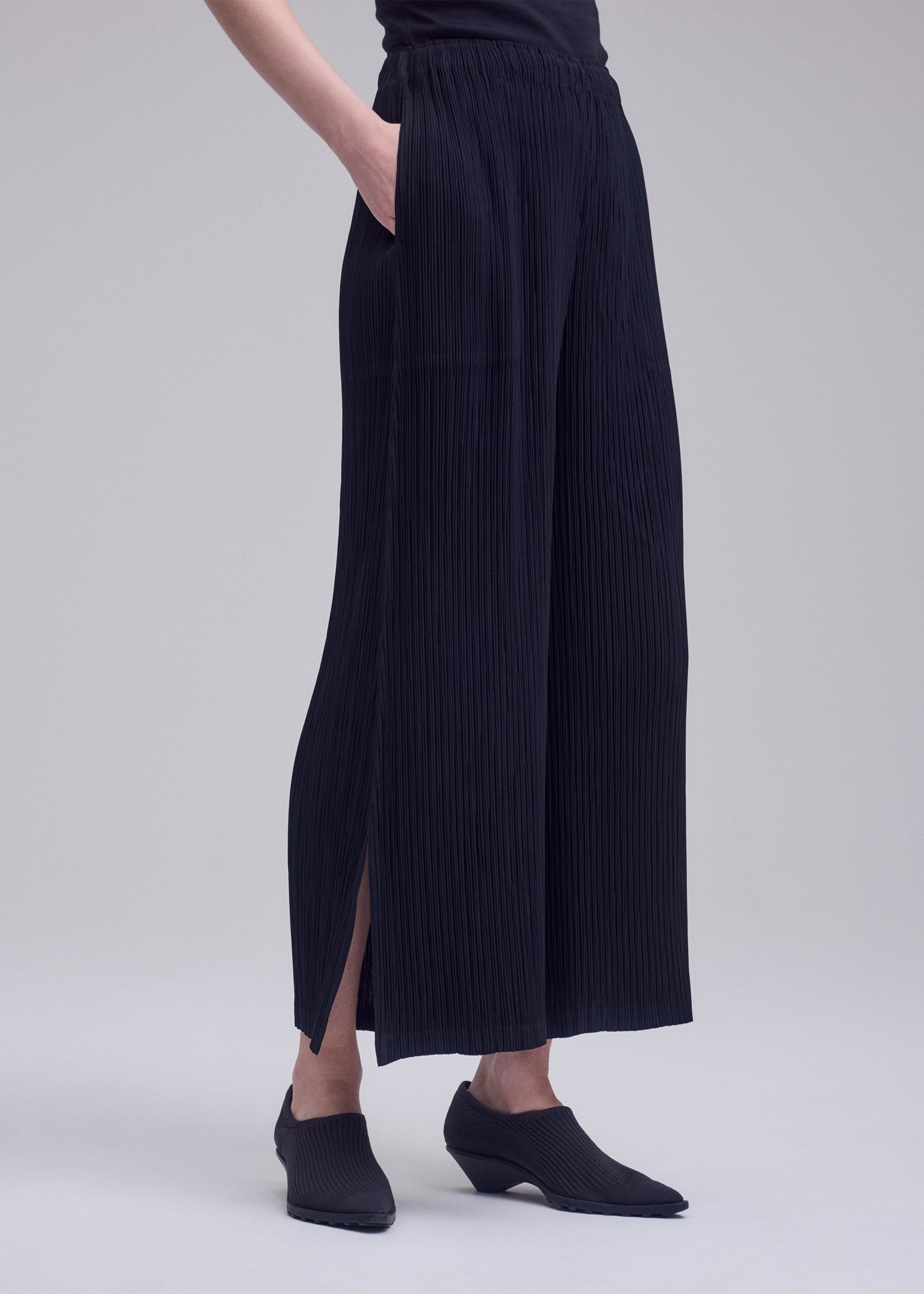 Zoomed view A person stands against a plain light gray background, wearing ISSEY MIYAKE’s HATCHING BOTTOMS—loose, pleated wide-leg pants with a textured finish and subtle side slits crafted from soft polyester—paired with black pointed shoes.