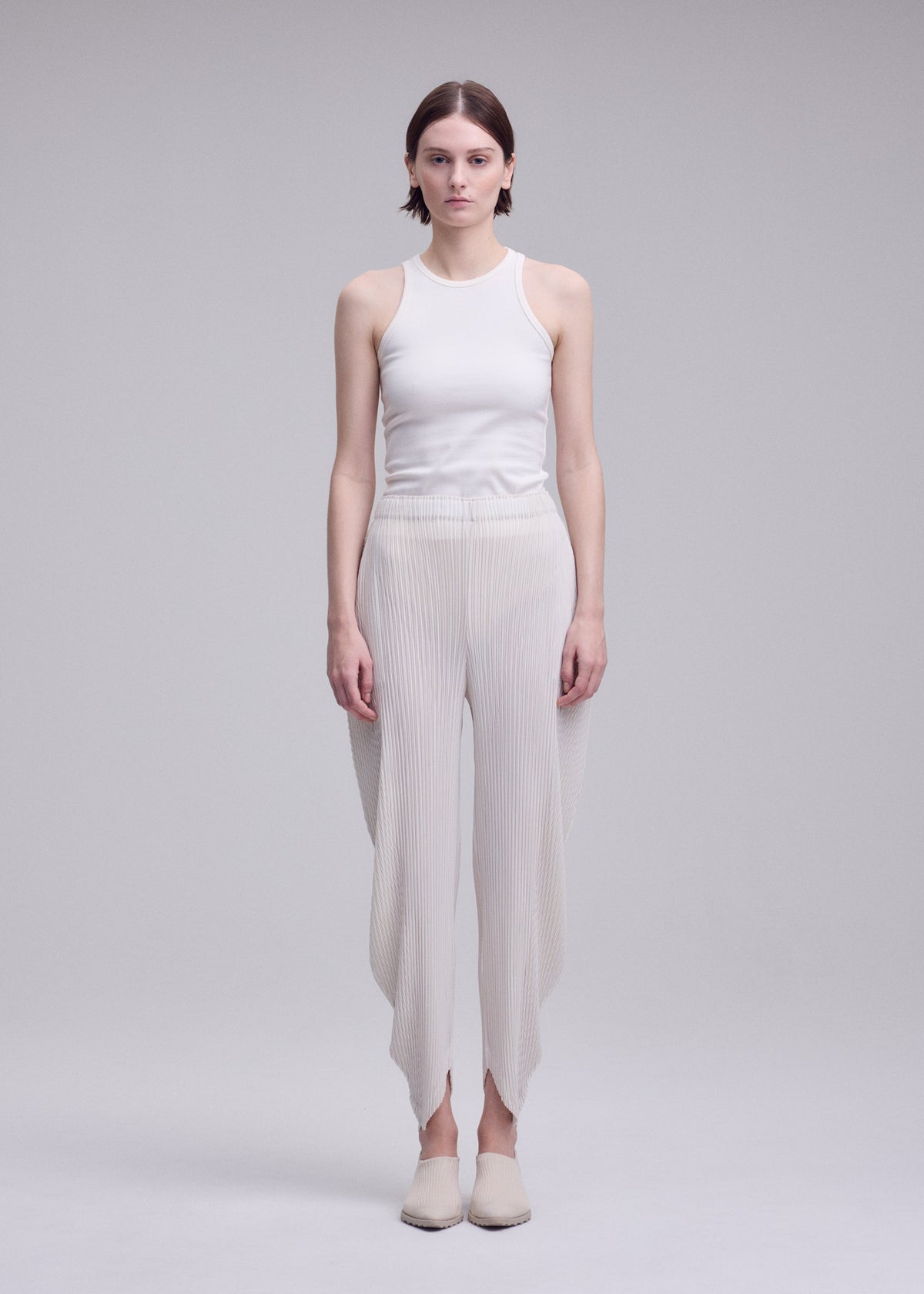 A woman stands facing forward against a plain light gray background, wearing the ISSEY MIYAKE LEAF PLEATS: a sleeveless white top paired with double-pleated, asymmetrical pants, and flat shoes. view 6 of 8