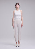 A woman stands facing forward against a plain light gray background, wearing the ISSEY MIYAKE LEAF PLEATS: a sleeveless white top paired with double-pleated, asymmetrical pants, and flat shoes.